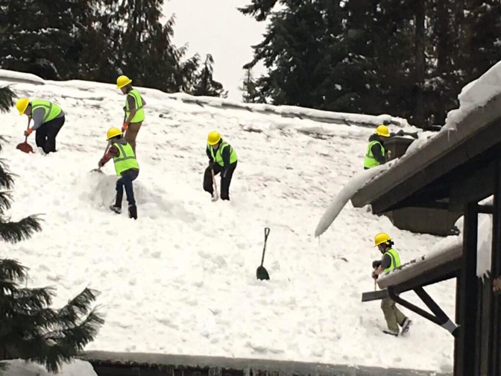 Professional crew wearing hard hats and safety gear safely clearing snow from a residential roof in Priest Lake.